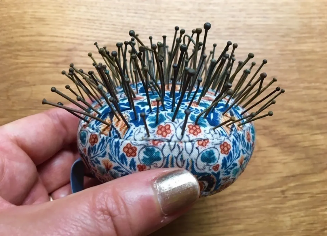 A hand holding a fabric pincushion with a floral pattern, filled with many straight pins and needles sticking out in various directions.
