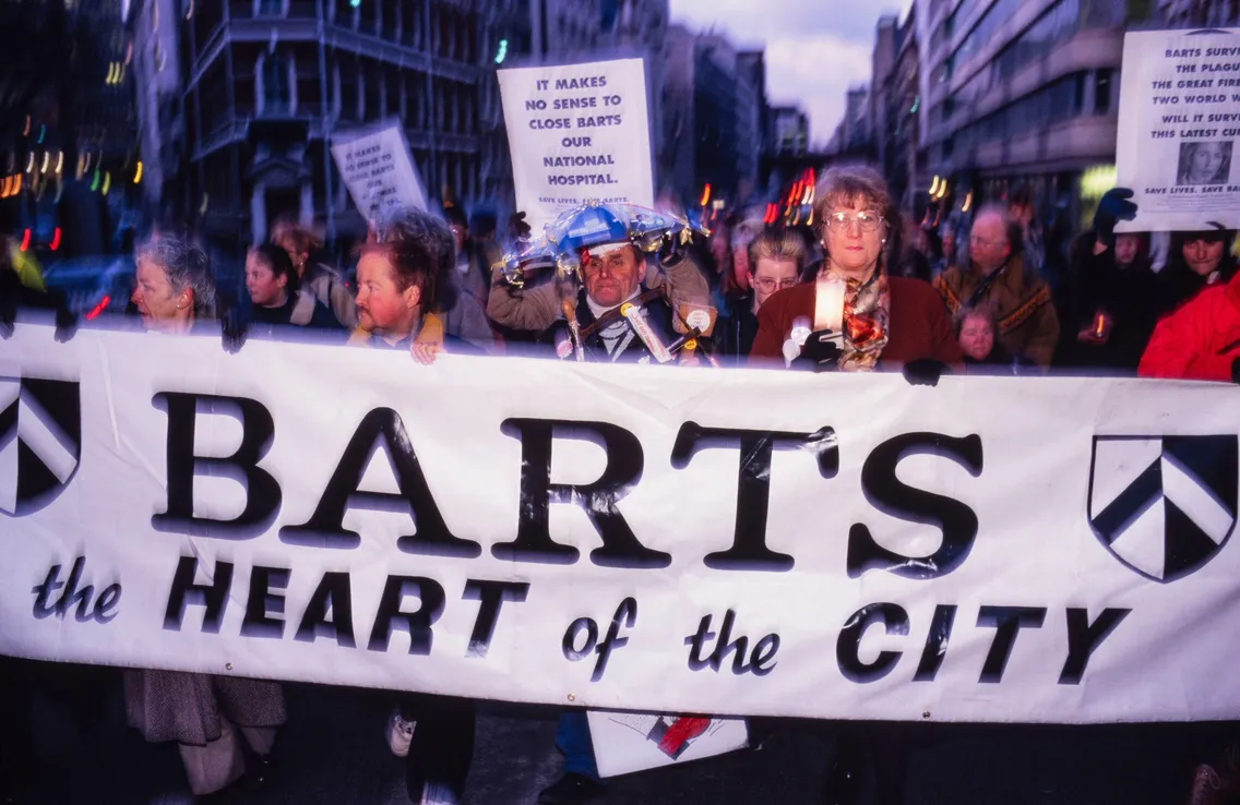 A group of people march on a city street holding a large banner reading "Barts the Heart of the City" to protest the hospital's closure, with additional signs visible in the background.