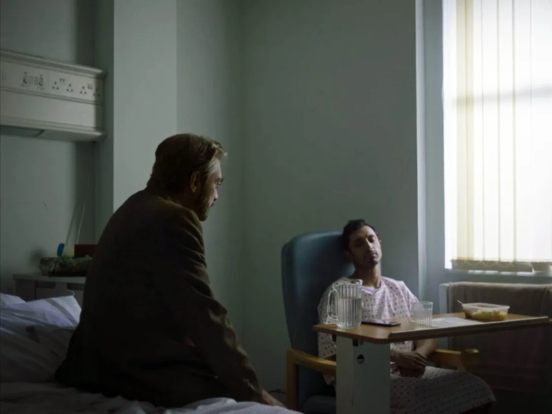 A man in a hospital gown sits in a chair beside a bed, talking to another man in a suit in a dimly lit hospital room.