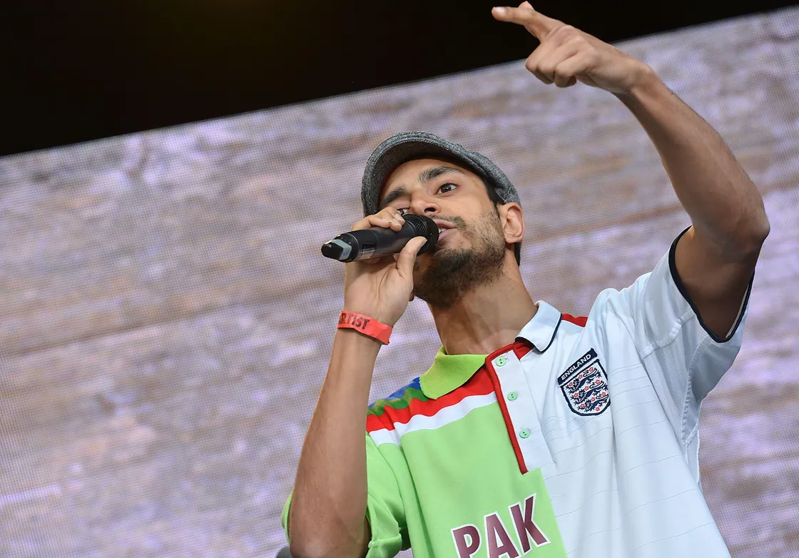A man wearing a cap and a colorful shirt speaks into a microphone on stage, gesturing with his hand.