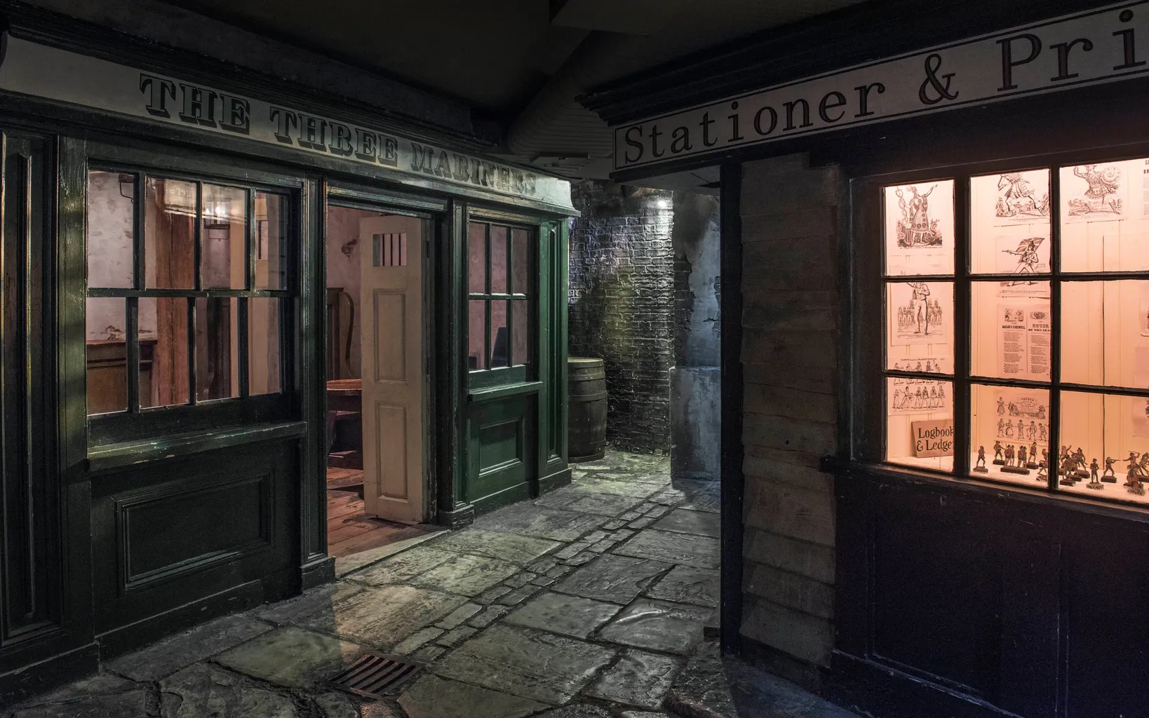Dimly lit alley with a vintage pub, "The Three Mariners," and a stationer's shop. Illuminated windows reveal interior details; cobblestone path leads between buildings.