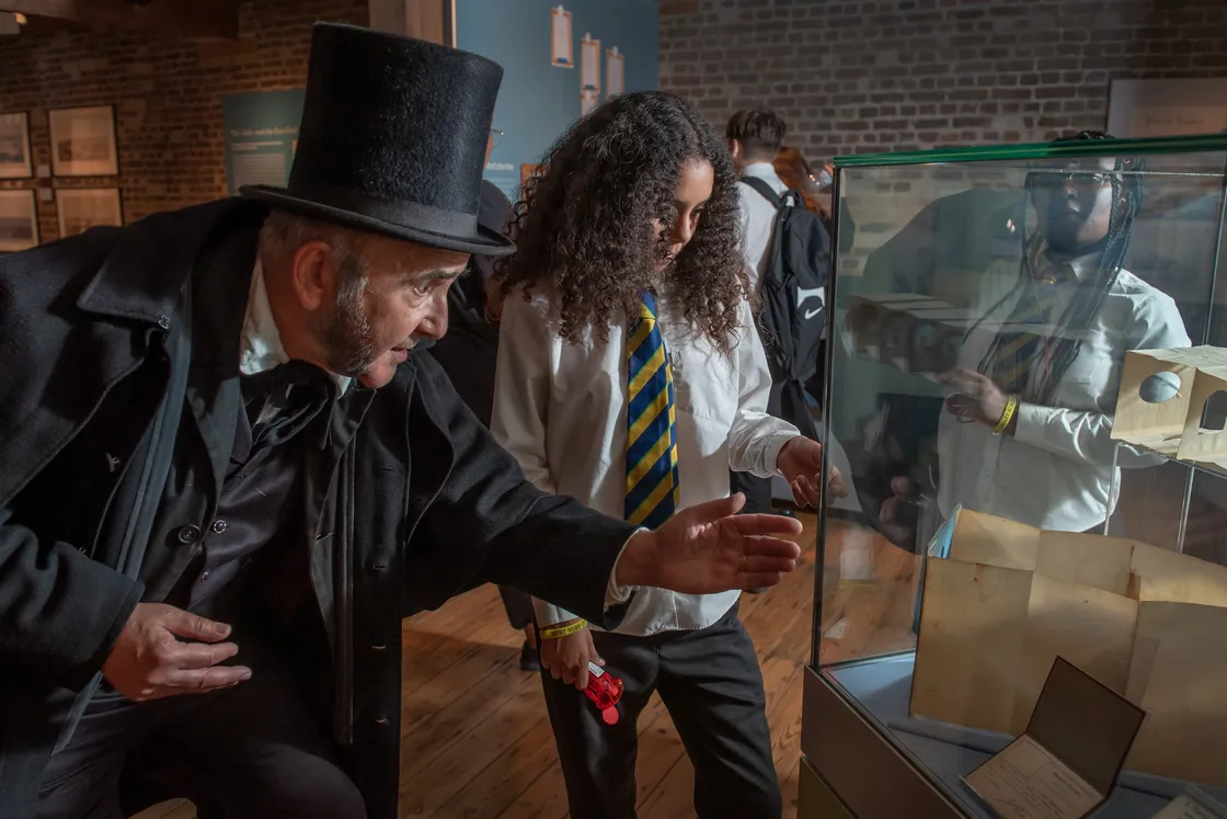 A man in period costume and a student in school uniform examine historical documents in a museum display case.