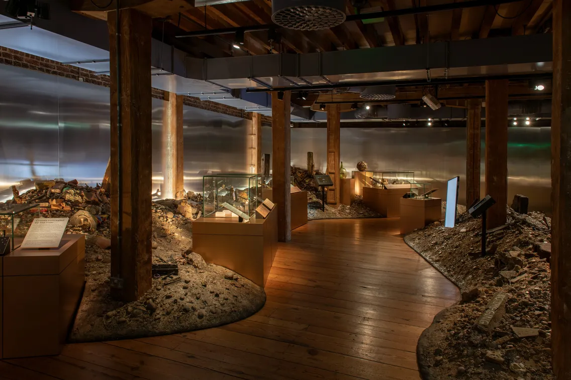 A museum exhibit room with wooden floors, columns, and display cases featuring artifacts and debris on mounds, under dim lighting.
