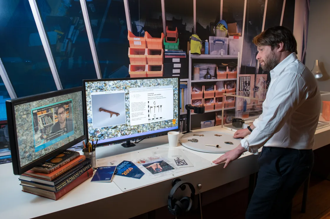 A person stands at a desk with two computer monitors, books, headphones, and various objects scattered around in a room filled with shelves and tools.