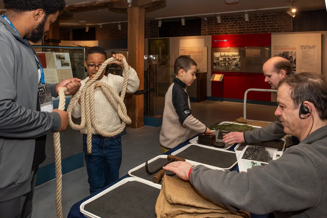 Children and adults interact with historical artifacts, including ropes, at a museum exhibit.