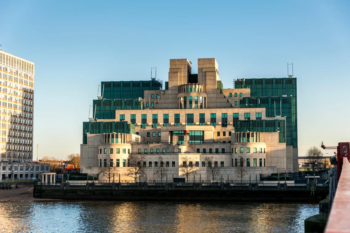 A large, modern building with green and beige sections sits beside a river under a clear blue sky.