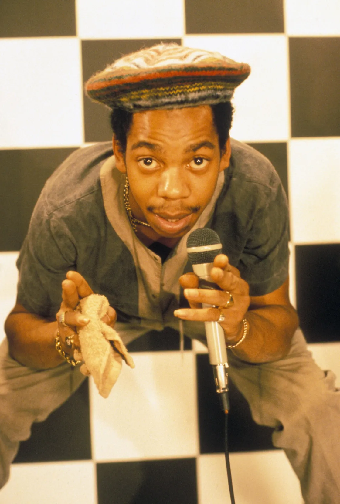 A man wearing a striped cap and gray outfit holds a microphone and a cloth, posing in front of a black and white checkered background.