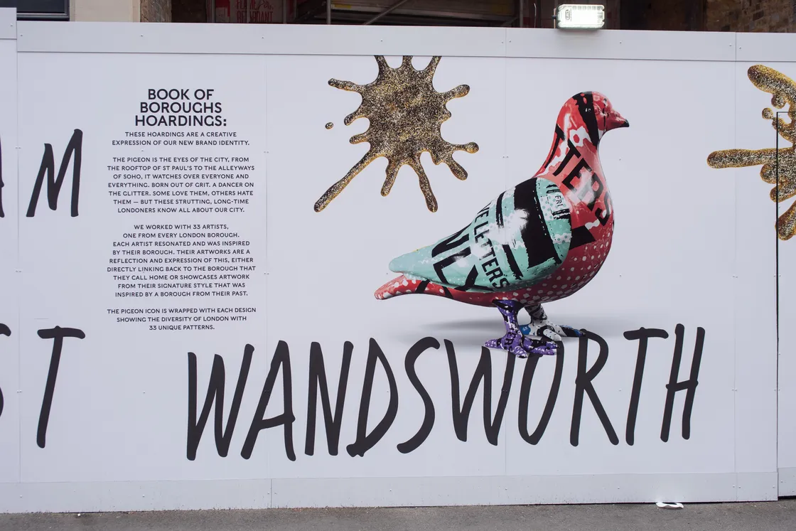 A colorful pigeon sculpture with mixed text and patterns is displayed on a white wall with black lettering reading "WANDSWORTH" and a gold paint splatter design.