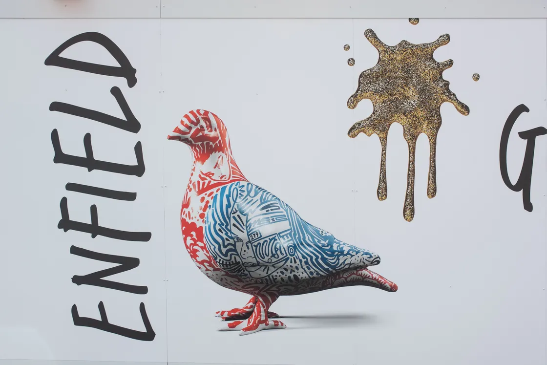 A pigeon statue with red, white, and blue patterns is displayed on a white background beside the word "ENFIELD" and a gold paint splatter.