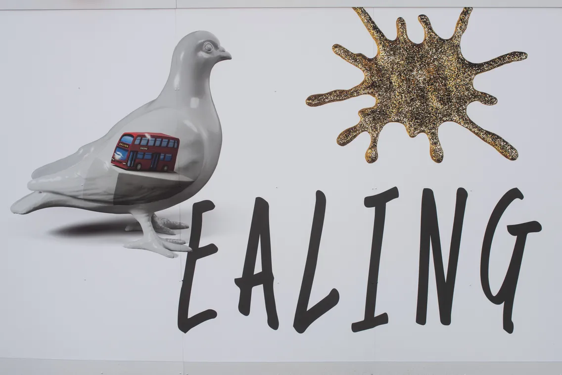 A graphic of a grey pigeon with a red double-decker bus inside it, next to a gold splatter shape and the word "EALING" in bold black letters.
