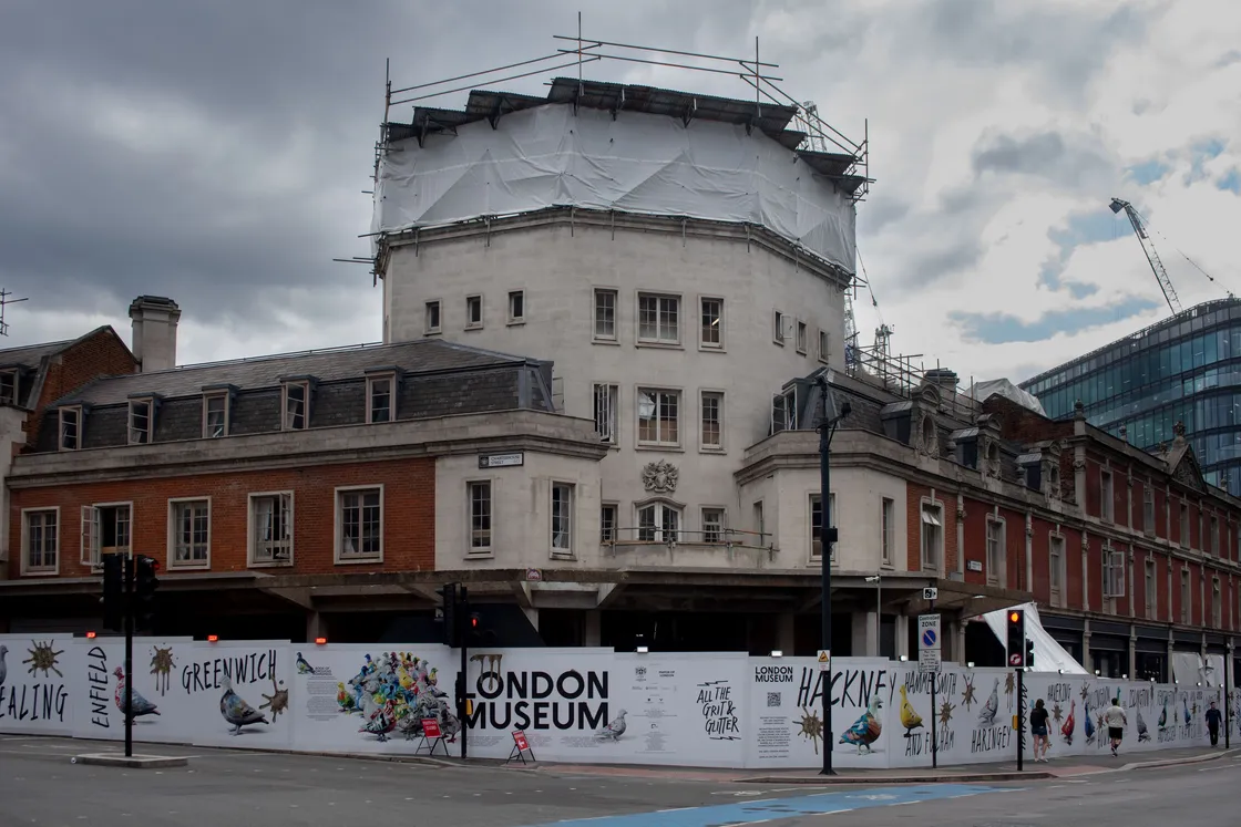 A large building, the Smithfield Market, under renovation is covered in scaffolding and white sheeting, with construction barriers displaying "London Museum" and neighborhood names at street level.