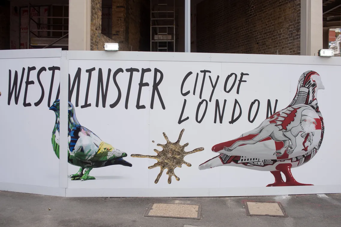 A construction wall mural features the text "Westminster City of London" with two painted pigeons and a gold splatter in the center.