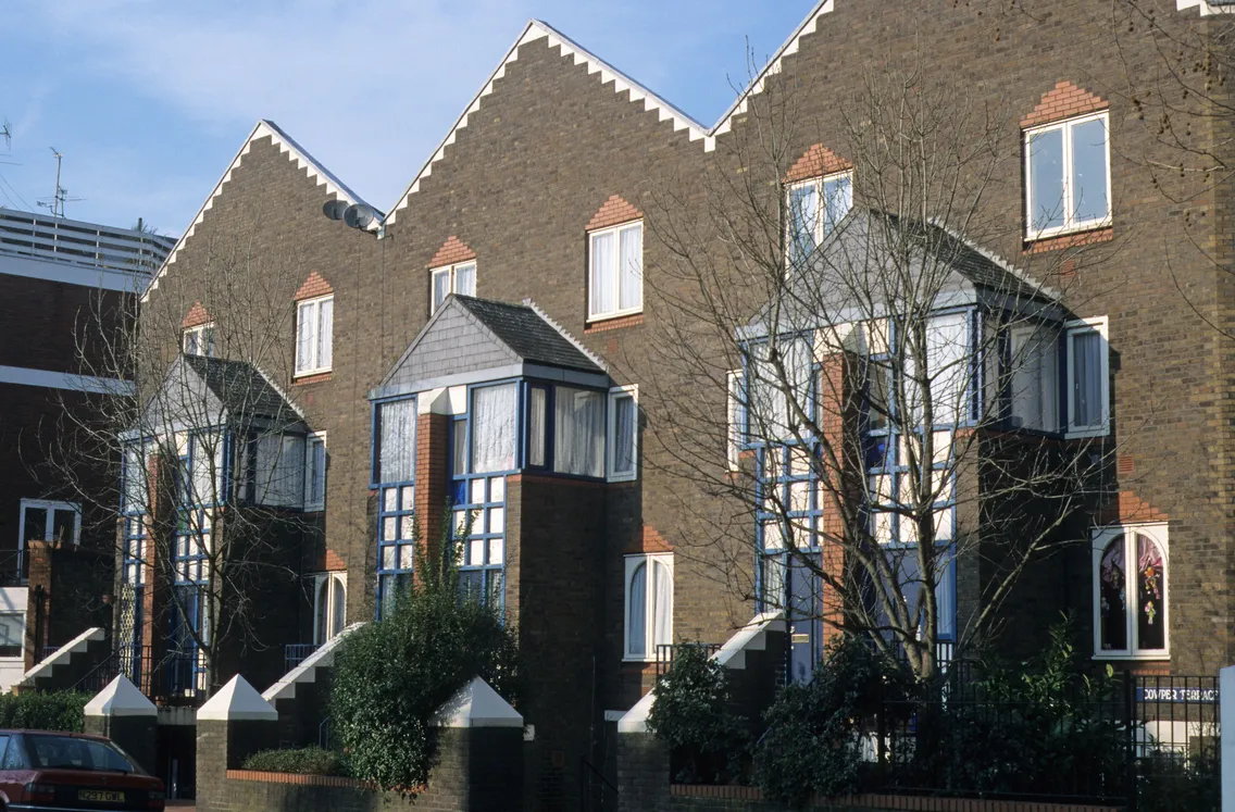 Row of modern brick townhouses with steep gabled roofs, geometric windows, and bare trees in front, photographed during the day.
