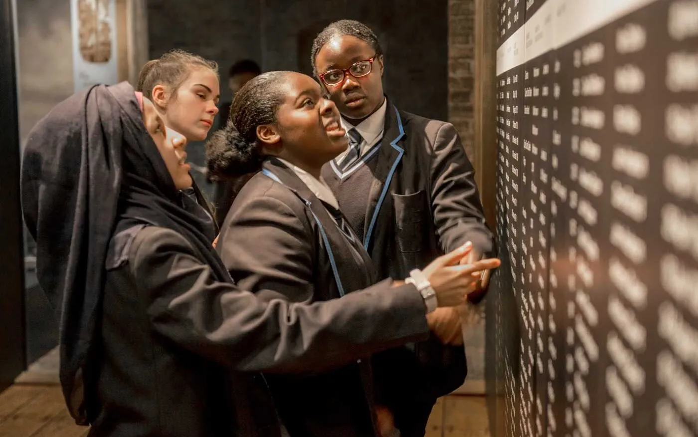 Four students in school uniforms examine a large wall display with numerous names. One student is pointing at the wall while others observe closely.
