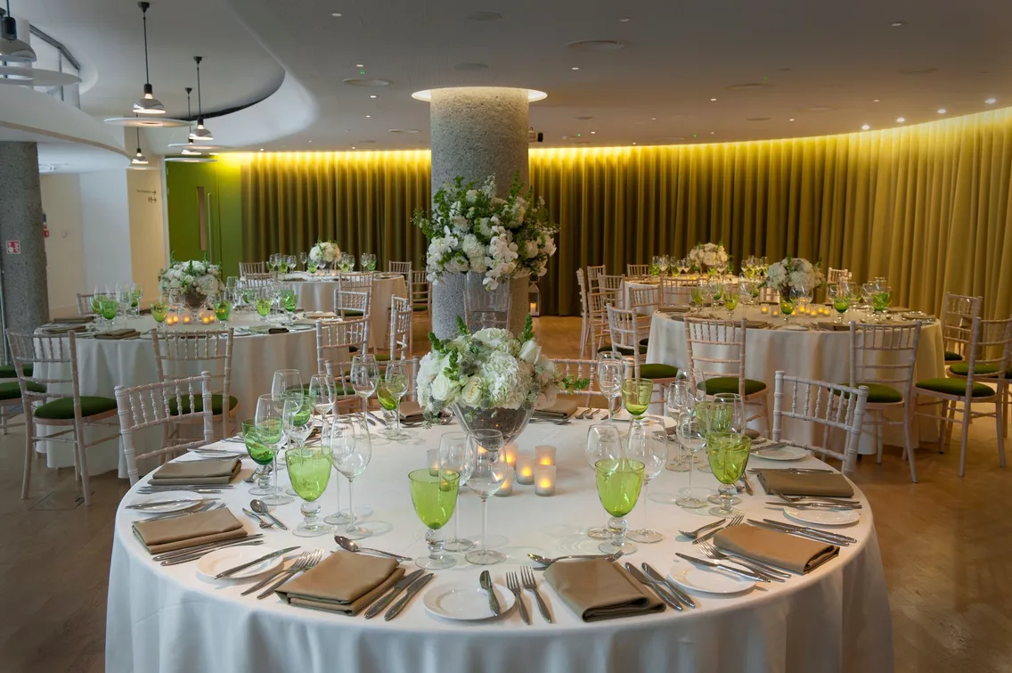 A banquet hall is set for an event with round tables covered in white tablecloths, navy blue napkins, and elegant glassware. Large windows and striped wooden paneling are in the background.