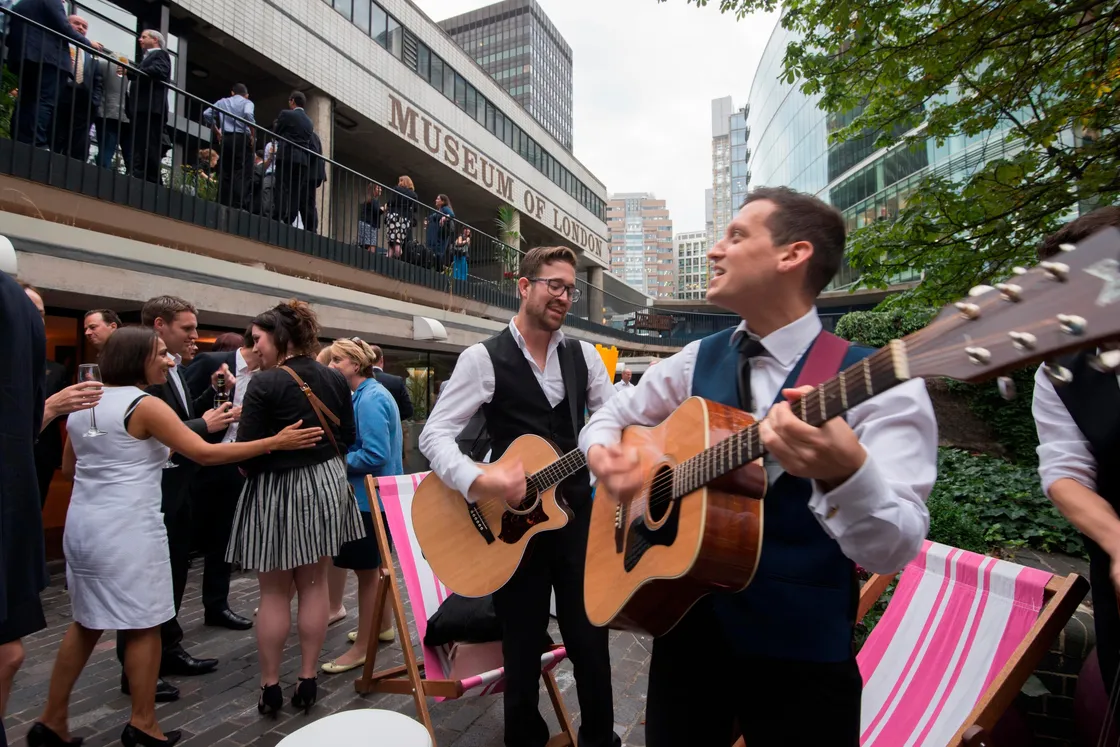 Two musicians play guitars in an outdoor space near the Museum of London. People socialize, some sitting on deck chairs. Surroundings include tall buildings and greenery.