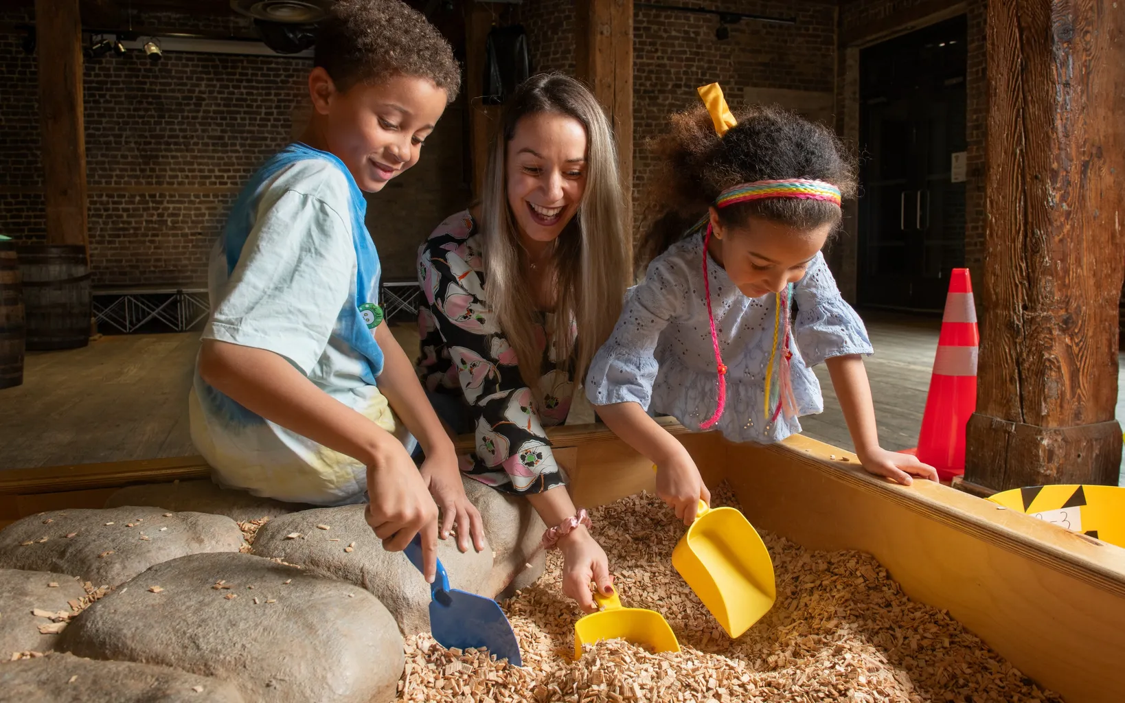 An adult and two children use plastic shovels to dig in a wooden sensory bin filled with wood chips, inside a room with brick walls and wooden beams.