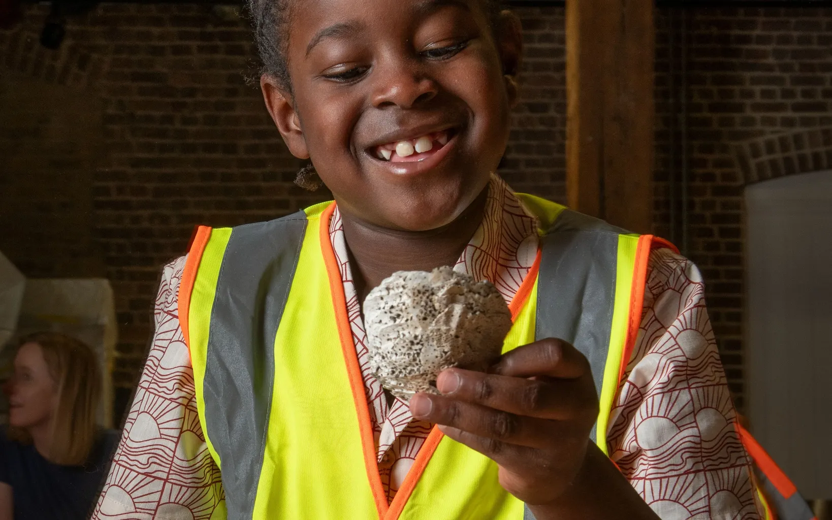 A child wearing a high-visibility vest smiles while holding a rock and a yellow scoop, standing indoors with exposed brick and beams in the background.