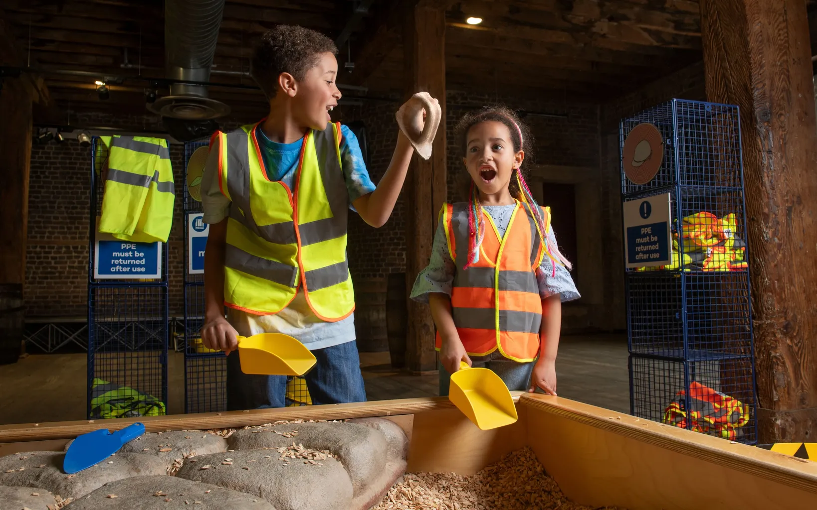Two children in  high vis jackets stand by a trench. Both are excitedly looking at a discovered artefact.