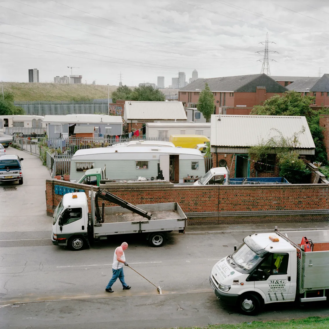 A worker sweeps a street near parked cars and trucks, with industrial buildings and a city skyline in the background.