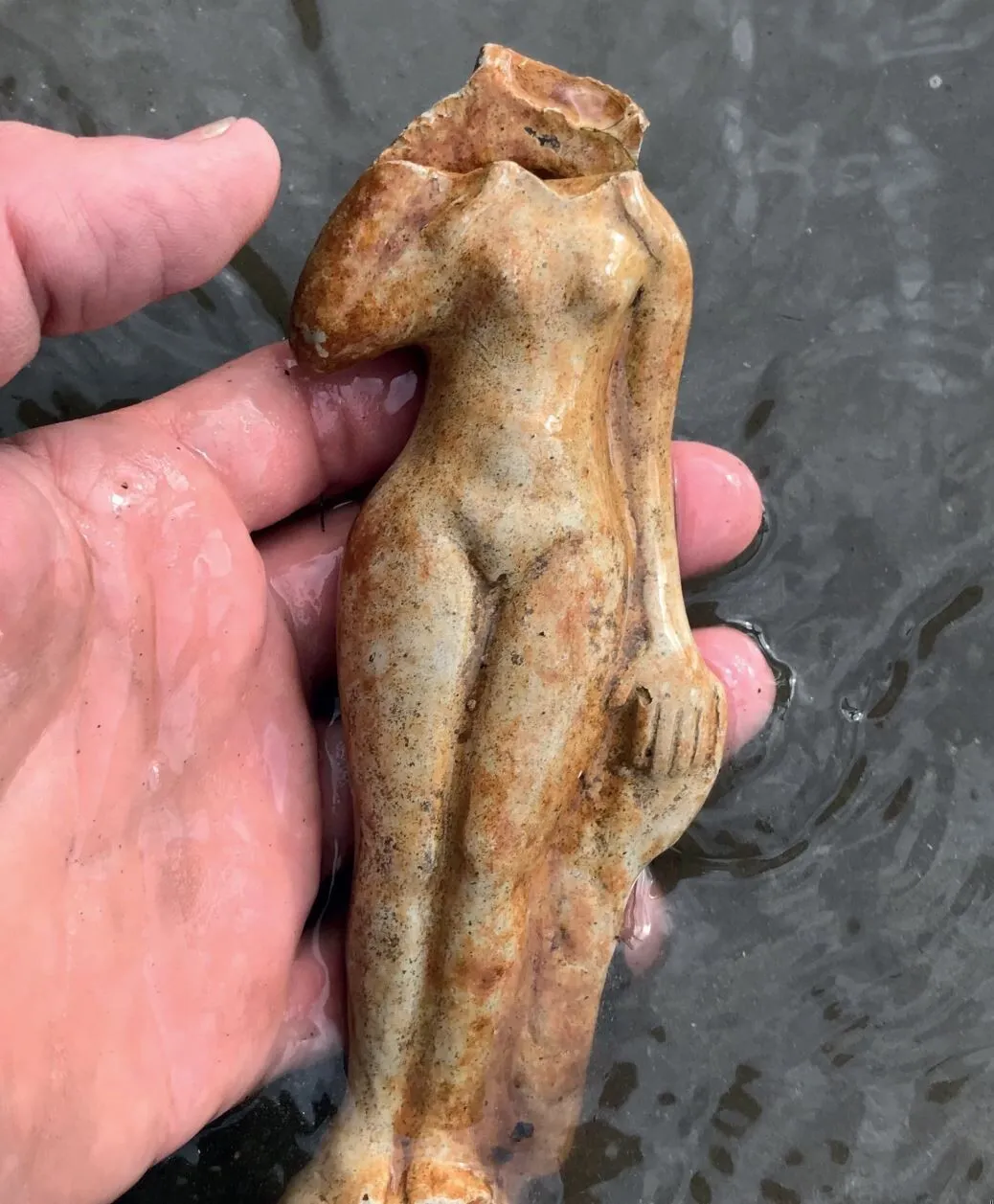 A hand holds a weathered, headless ceramic figurine of a nude woman, in the background of flowing water.