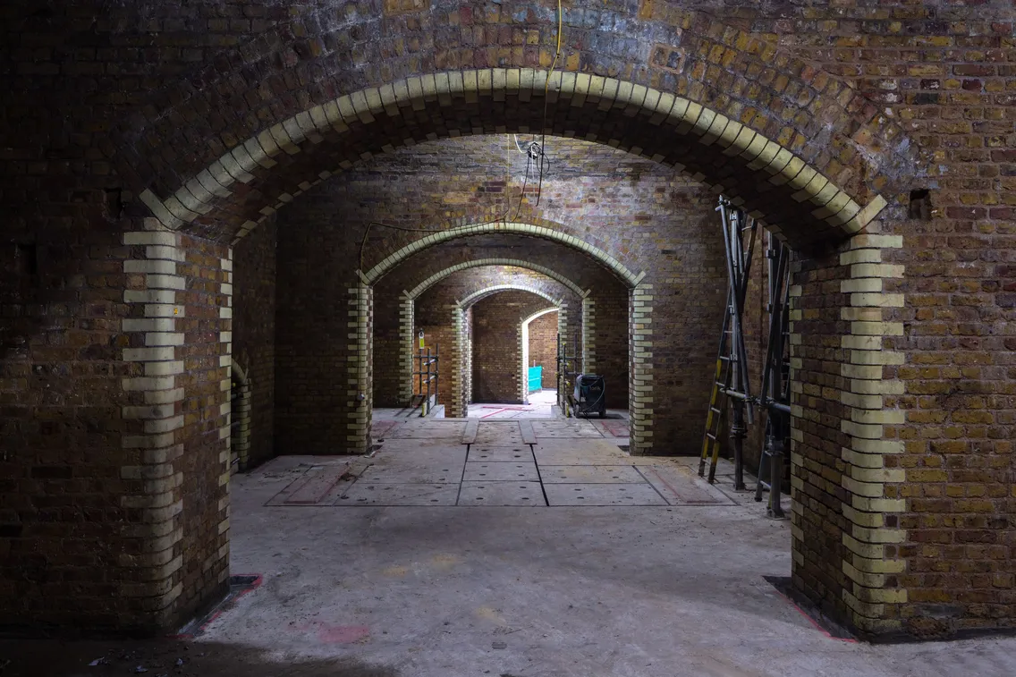 A dimly lit underground brick tunnel with multiple archways. Construction materials and equipment are visible in the background.
