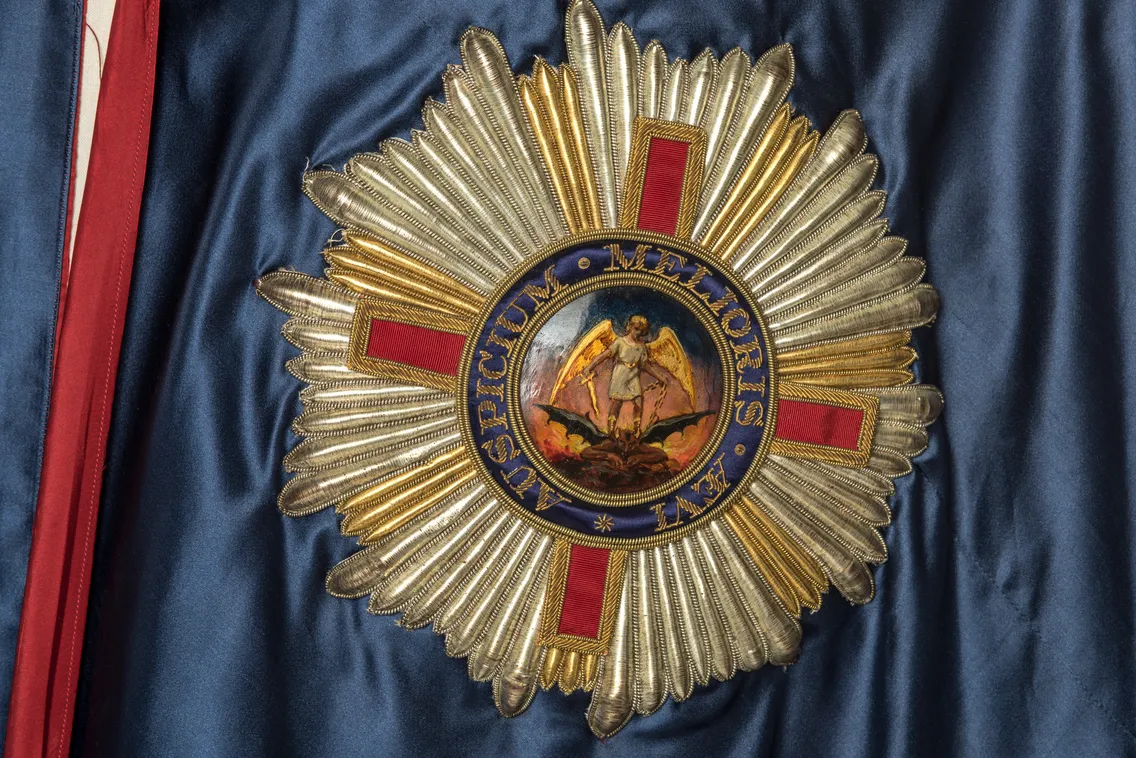 A detailed emblem featuring a gold starburst and a central medallion with a classical scene, set against a draped blue fabric with a red striped edge.