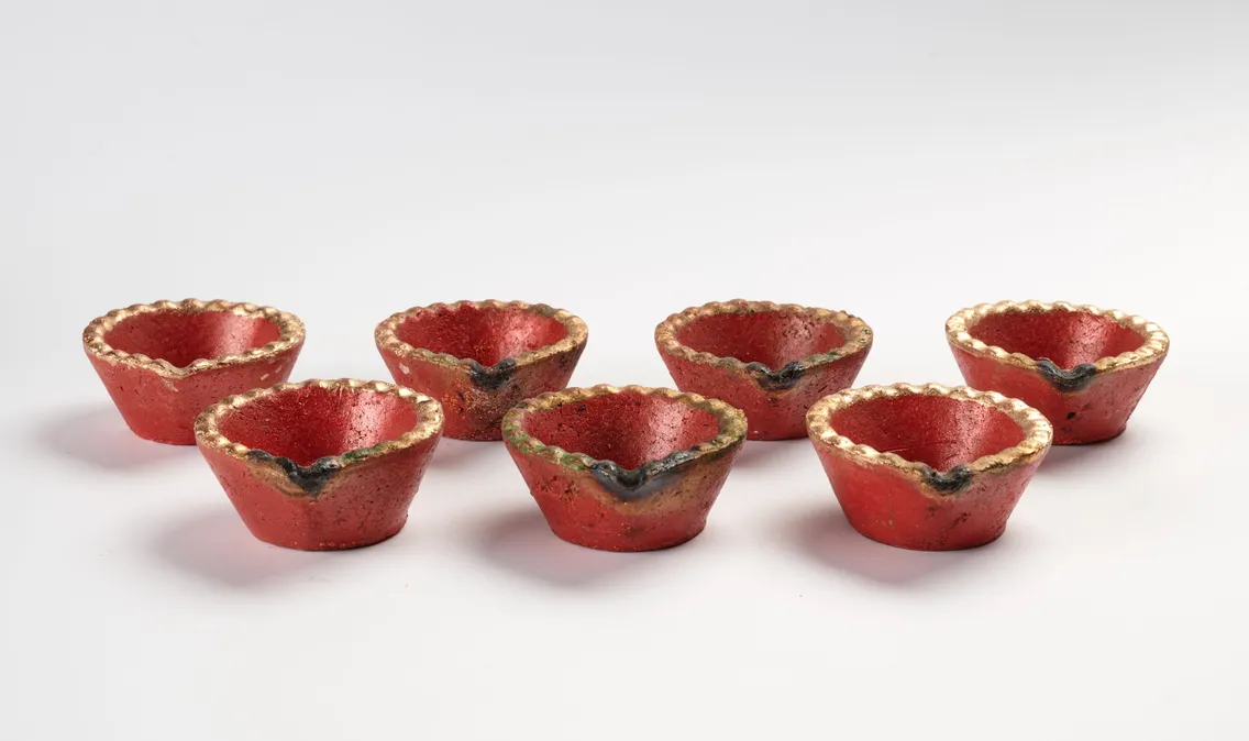 Seven small, red clay lamps with gold-edged rims arranged in a scattered formation on a white background.