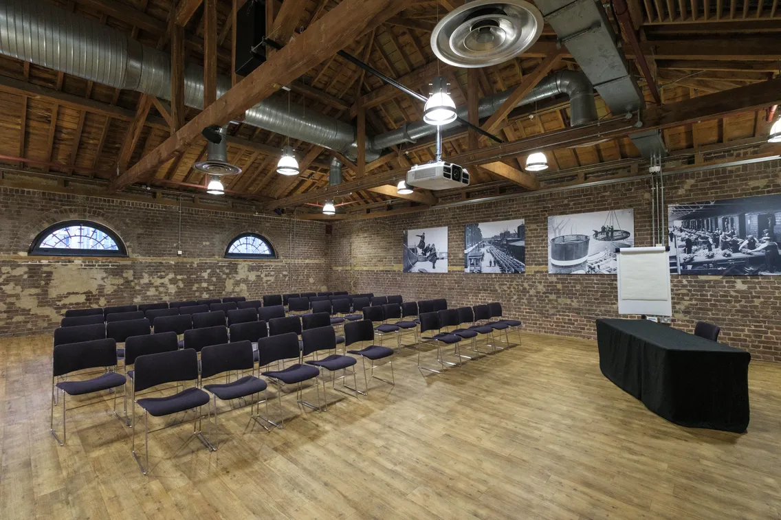 A conference room with rows of empty chairs, a projector, a screen, a flip chart, and a long table with a black tablecloth under exposed wooden beams and industrial ducts. Black-and-white photos adorn the walls.