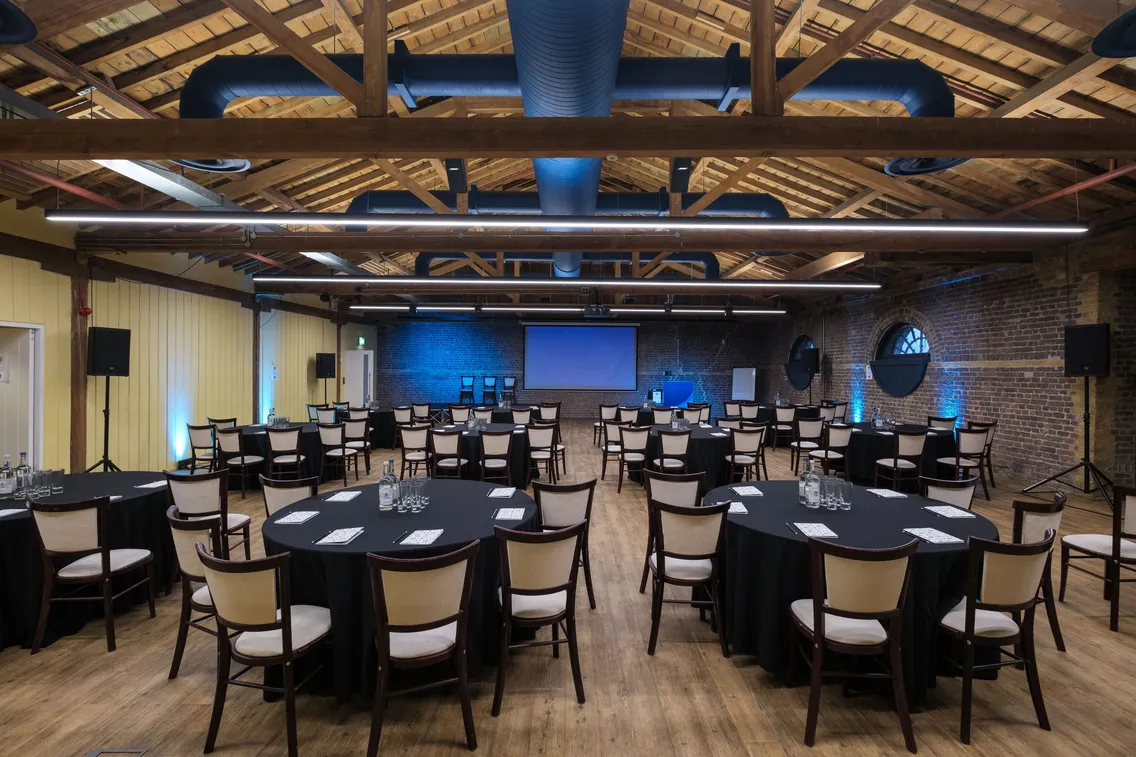 A conference room with round tables and chairs arranged facing a large screen. The room has exposed wooden beams and brick walls, with blue lighting and various equipment set up.