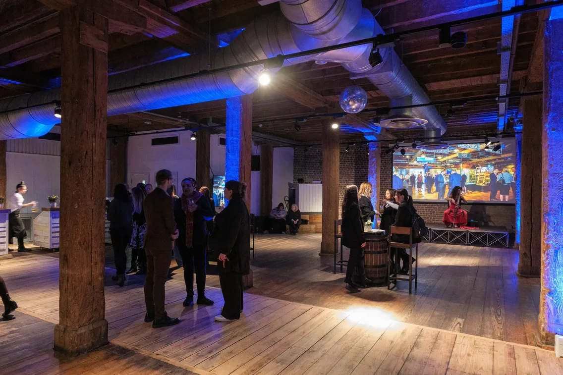 A group of people socialize and interact in a spacious, industrial-style room with wooden beams and flooring, featuring a large screen displaying colorful images in the background.