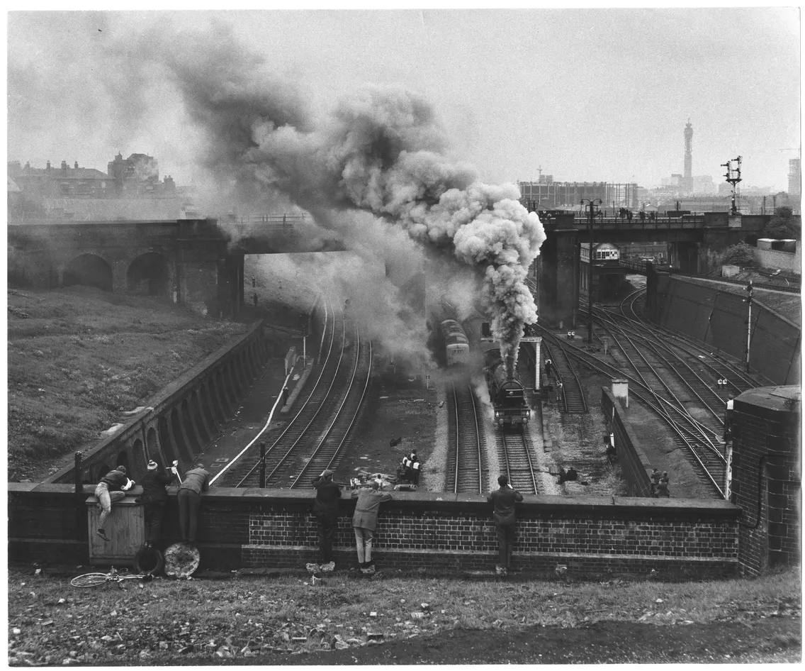 A vintage black-and-white photo of a steam train emitting thick smoke while traveling on railway tracks, with people watching from a nearby wall.
