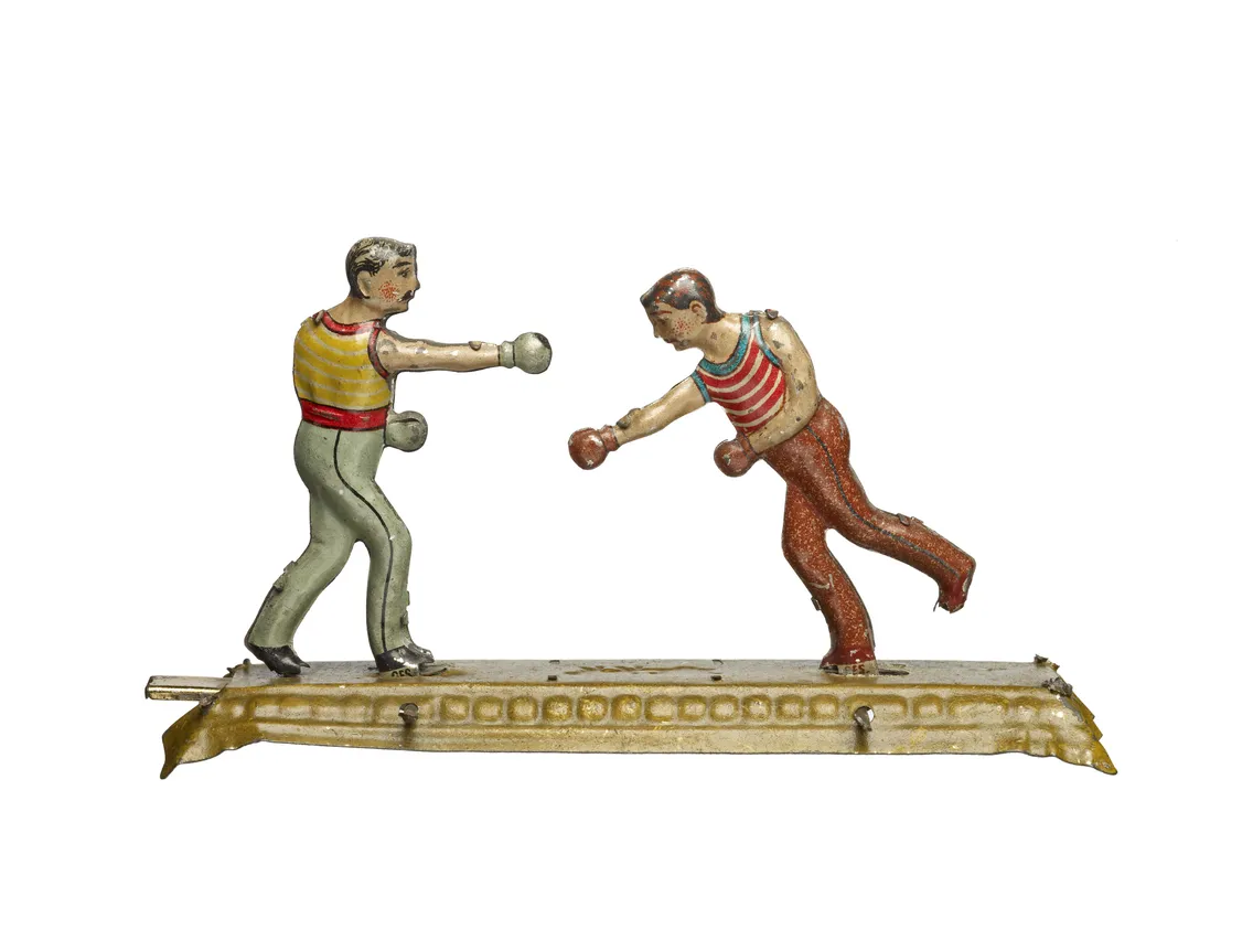 Two vintage toy boxers fighting on a metal base, one in red and the other in green pants, both wearing striped shirts.