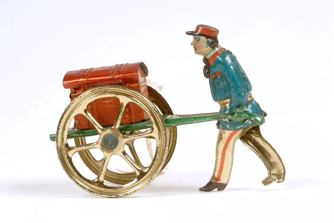 Vintage toy figure of a man pushing a red and gold fire hose reel on wheels, isolated on a white background.