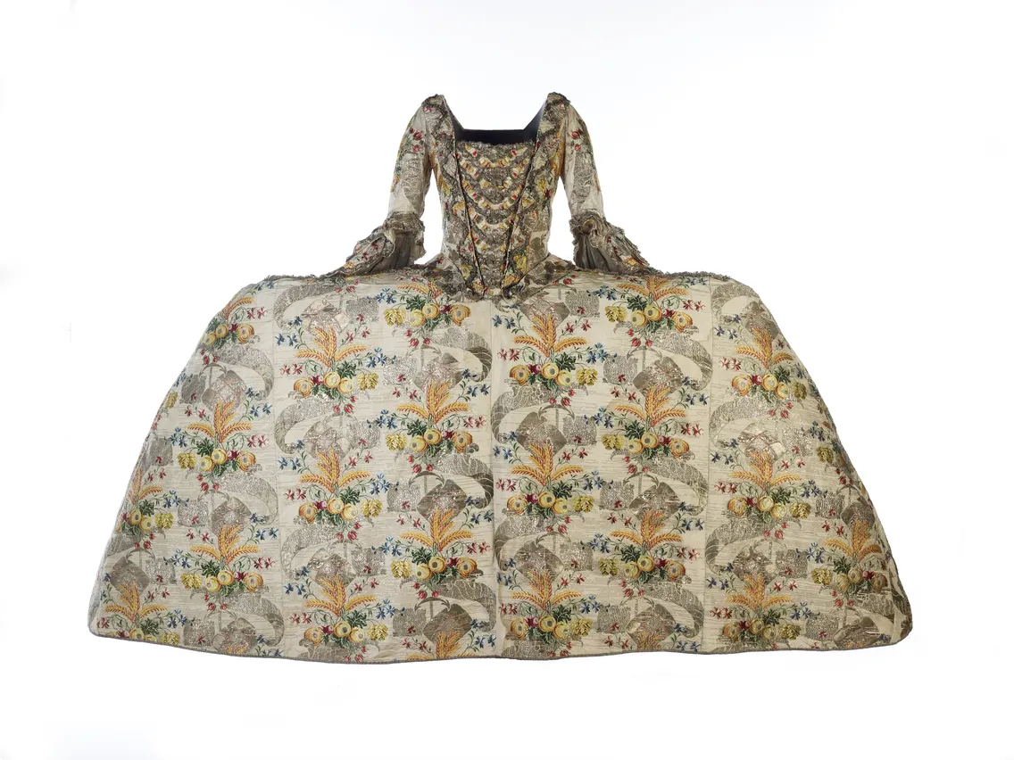 Historical dress from the 18th century with a wide hoop skirt, ornate floral and scroll embroidery, displayed on a white background.