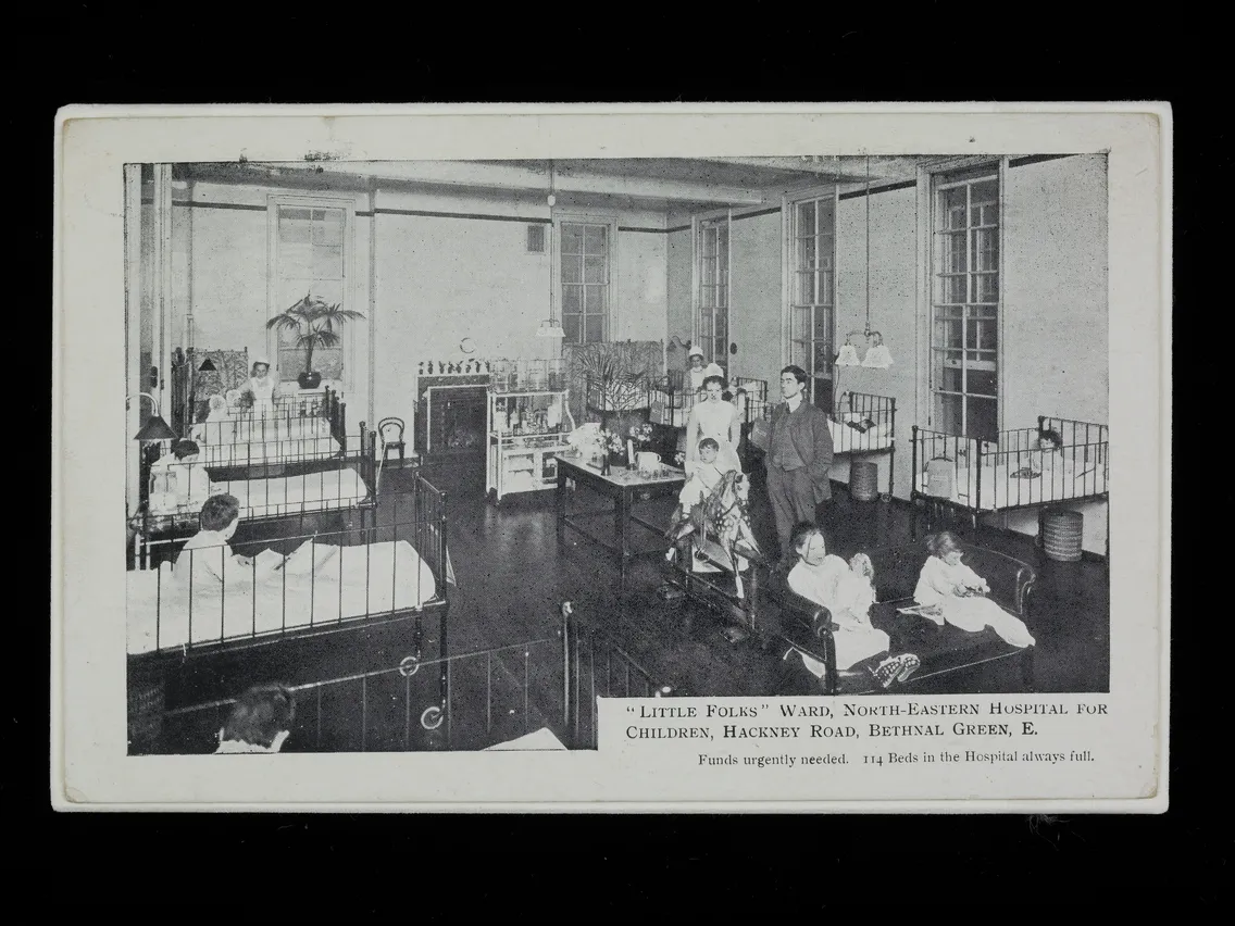 Black and white historical photo of children and nurses in the "little folks" ward at the north-eastern hospital for children, london. beds and sparse furnishings are visible.