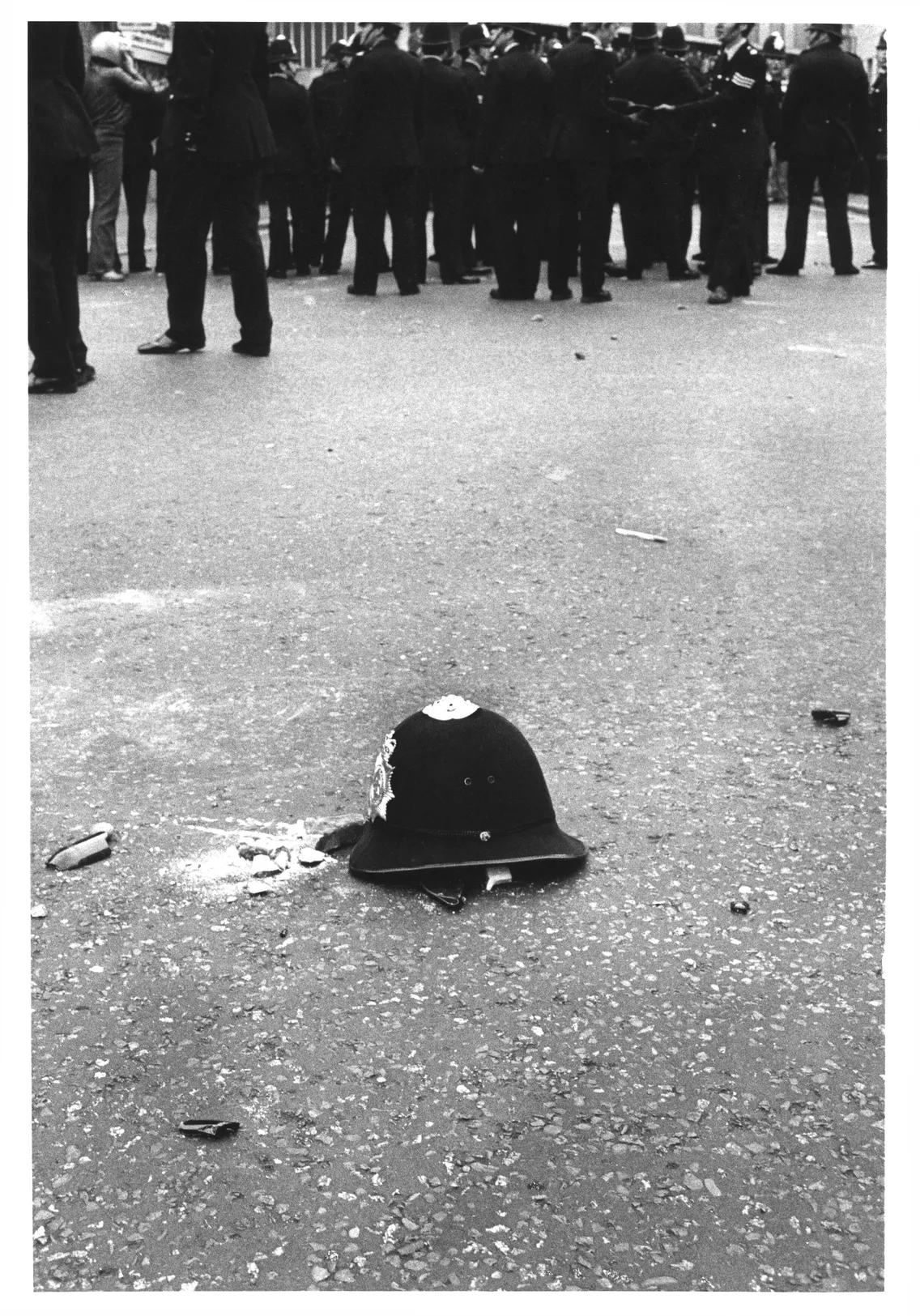 A police helmet lies in the foreground on a street scattered with debris, with a group of police officers and onlookers in the background.