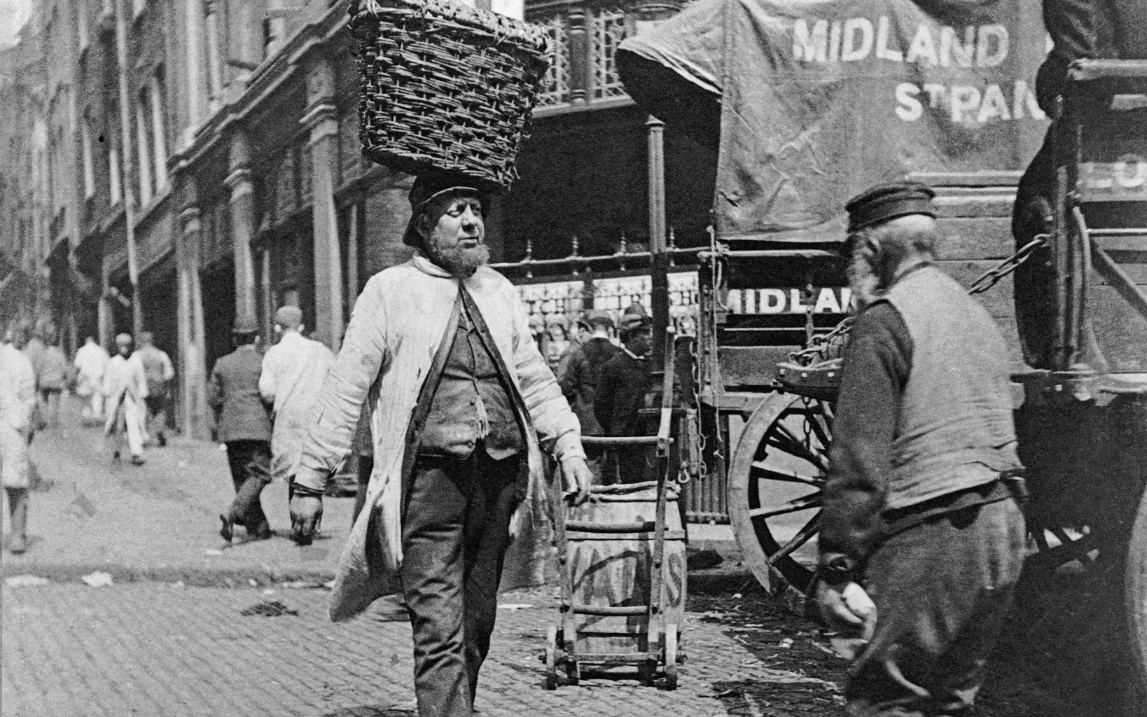 A historical black and white photo showing a man carrying a large basket on his head in a busy street with horse-drawn vehicles and pedestrians.