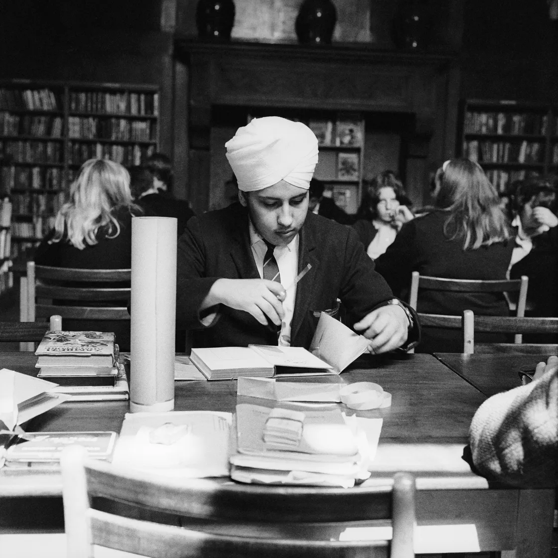 A young sikh man wearing a turban studying in a library, surrounded by books and other students.