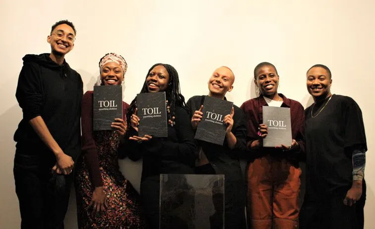 Six people stand in a row against a plain wall, with three of them holding copies of a book titled "TOIL." They are smiling and appear to be at a book launch or similar event.