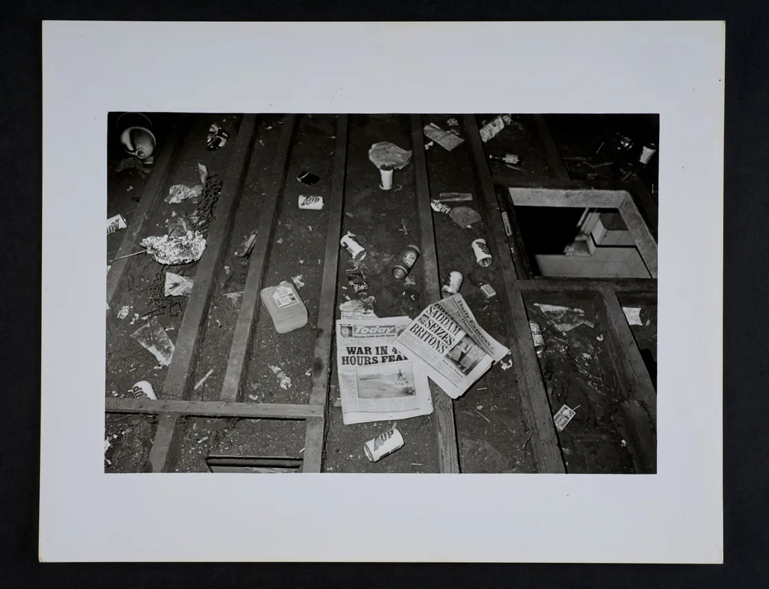 A black and white photo of a floor strewn with debris, papers, and a newspaper with the headline "War in 24 Hours?".