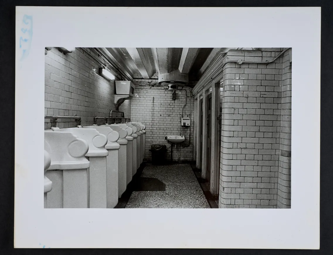 A black-and-white photograph of a public restroom with urinals on the left, a sink in the center, and stalls on the right. The space has tiled walls and floor, with exposed pipes on the ceiling.