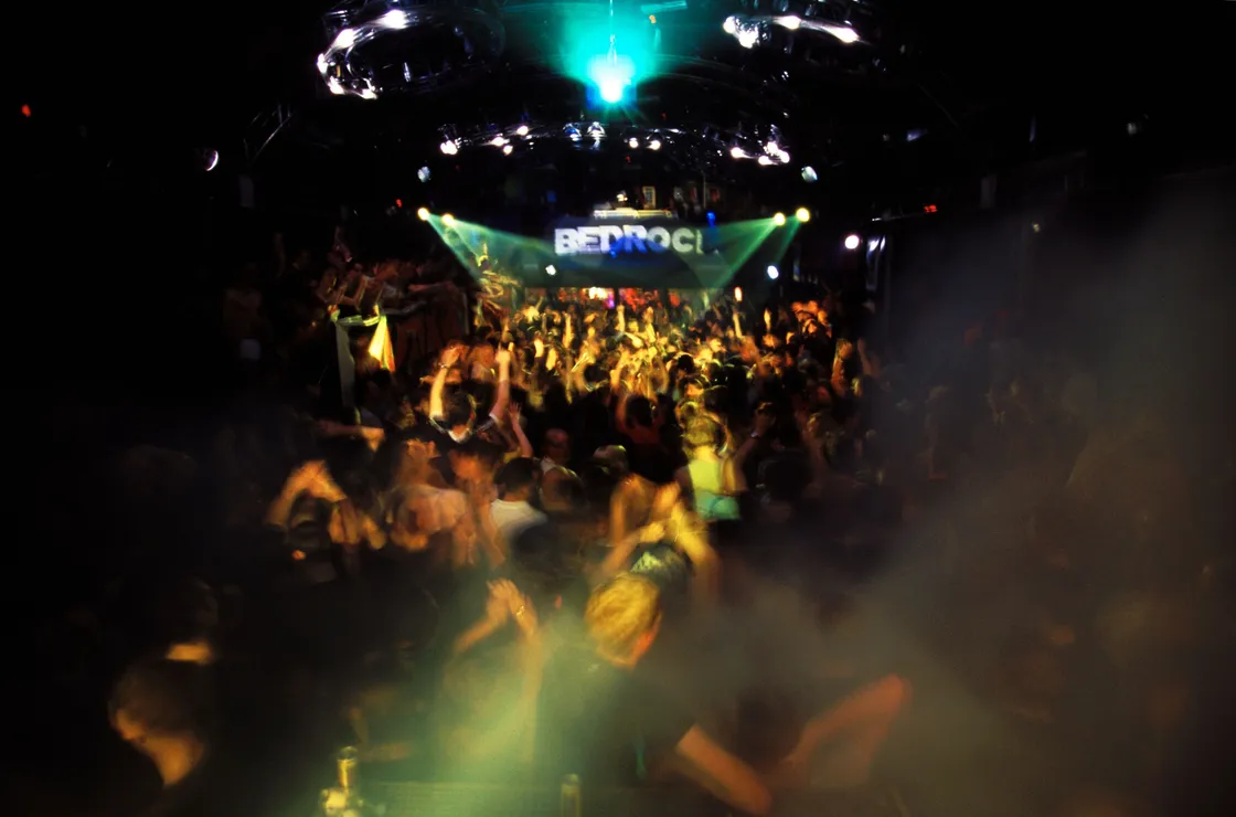 A dense crowd of people dance energetically in a dark nightclub with colorful lights and a DJ on stage. A sign reading "BEDROC!" is visible in the background.