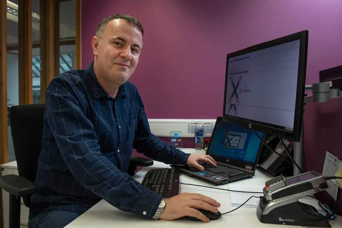 A man wearing a blue shirt with one hand on a computer mouse, the other on a computer keyboard. He is looking at the camera.