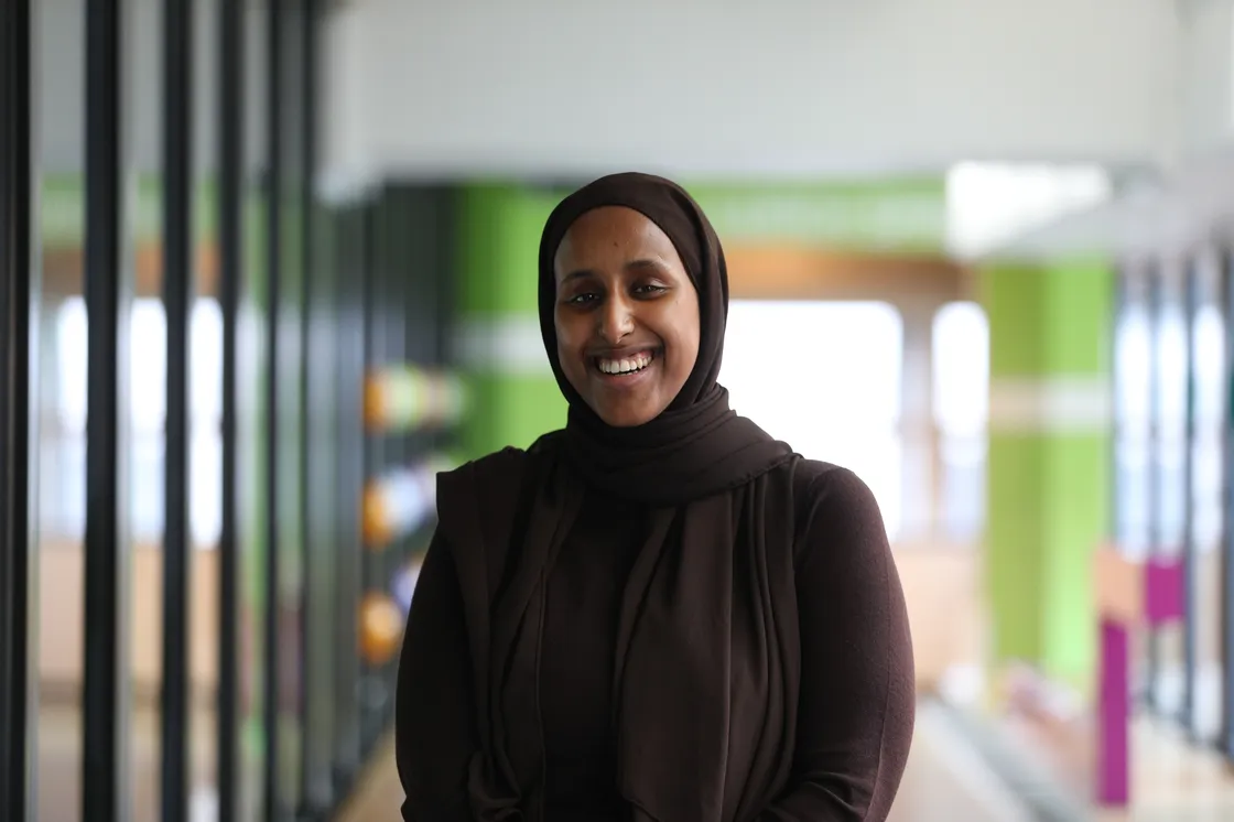 A smiling Black woman wearing a hijab standing in a corridor with green in the background.