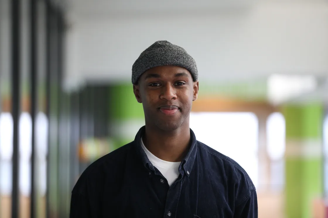 A smiling Black man wearing a grey hat in a corridor.
