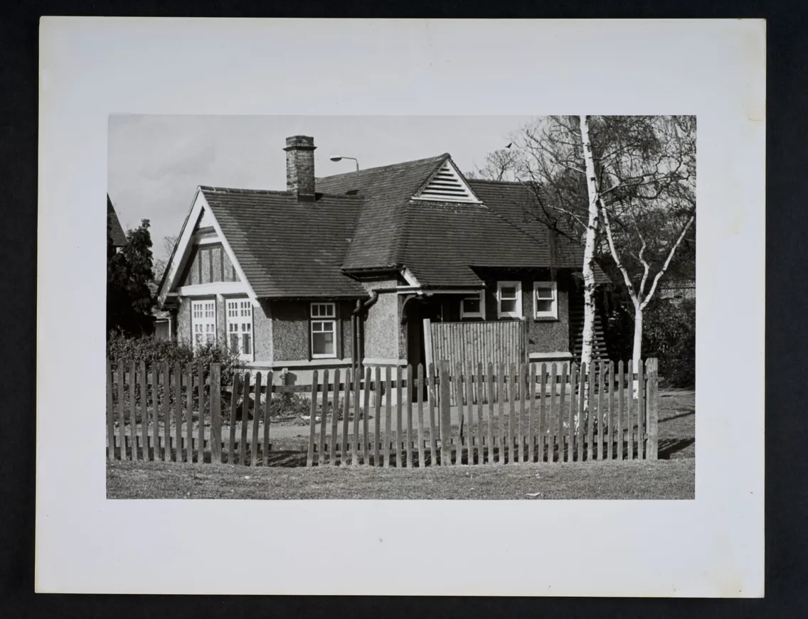Black and white photo of a small, single-story house with a slanted roof, fenced yard, and chimney, surrounded by trees.