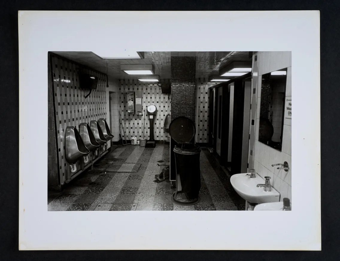 Black and white photo of a public restroom featuring urinals, a weighing scale, a sink, and multiple stalls, with a trash can in the center. Walls are tiled and flooring appears to be patterned.