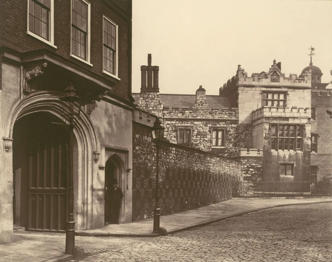 Sepia-toned vintage photo of a historic street corner with cobblestone pavement and traditional stone buildings, featuring an arched doorway and a gated fence.