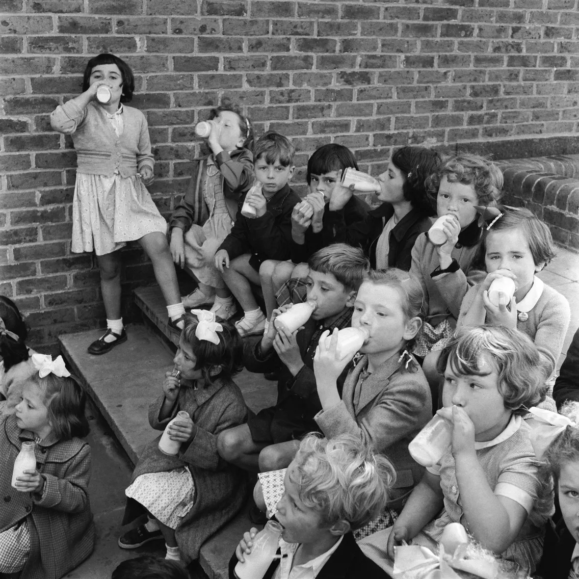 Children sitting on steps outside, drinking from glass bottles with straws. Some are looking at the camera, while others are focused on their drinks.